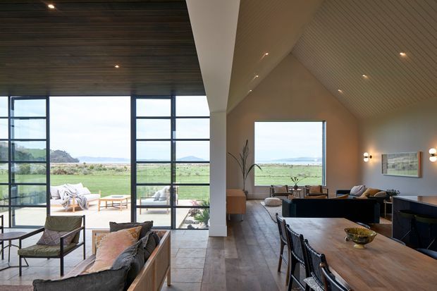 Open-plan contemporary living and dining area with a high pitched, timber-lined ceiling and expansive black-framed glazing overlooking a rural coastal landscape. The room includes a timber dining table with dark dining chairs, a lounge seating area, tiled flooring, recessed lighting, and a compact kitchen or bar zone at right. The interior is warm, refined, and strongly connected to the outdoors.