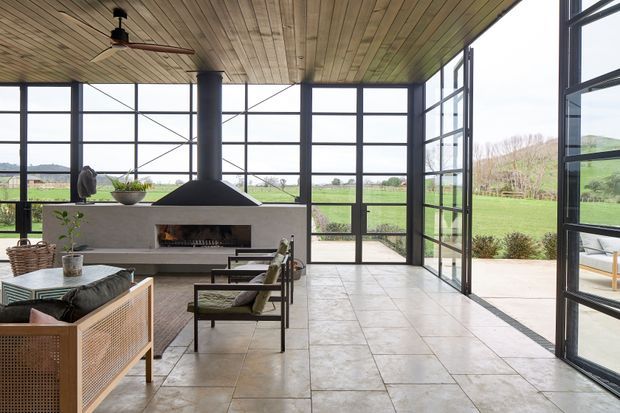 A bright, open-plan living room with floor-to-ceiling black-framed glazing overlooking a grassy rural landscape. The space features a suspended central fireplace with a broad hearth, timber-lined ceiling, large-format pale tiled flooring, woven-front seating, and understated mid-century-inspired furniture. A ceiling fan and extensive glazed walls reinforce the airy indoor-outdoor connection.