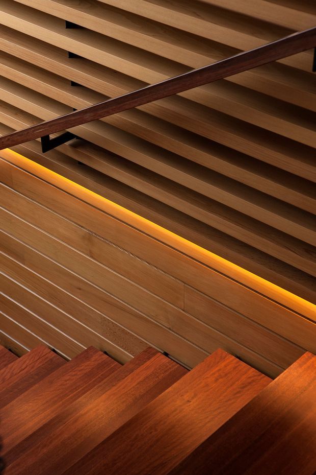 Close-up architectural view of a timber stairway, showing rich dark wood treads, a slatted timber ceiling or balustrade structure, a dark handrail, and warm concealed linear lighting integrated along the wall. The image focuses on material detail and layered horizontal lines rather than a wider room context.