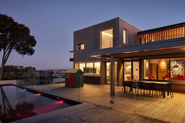 Twilight exterior view of a contemporary multi-level home with a box-like timber-clad upper volume, extensive glazing, and a large covered outdoor dining terrace. The timber deck surrounds a reflecting pool with illuminated accents, while a glazed balustrade and slatted roof structure frame the entertaining area. A residential neighbourhood and mature tree are visible in the background.