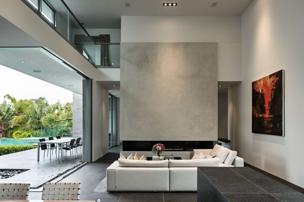 A sophisticated double-height contemporary living room with an open mezzanine and extensive floor-to-ceiling glazing. A large pale sectional sofa faces a dramatic concrete feature wall and fireplace area, while a dark stone-clad low table or hearth occupies the foreground. The room opens to an outdoor dining and pool area, and an abstract artwork adds a warm red and orange accent.
