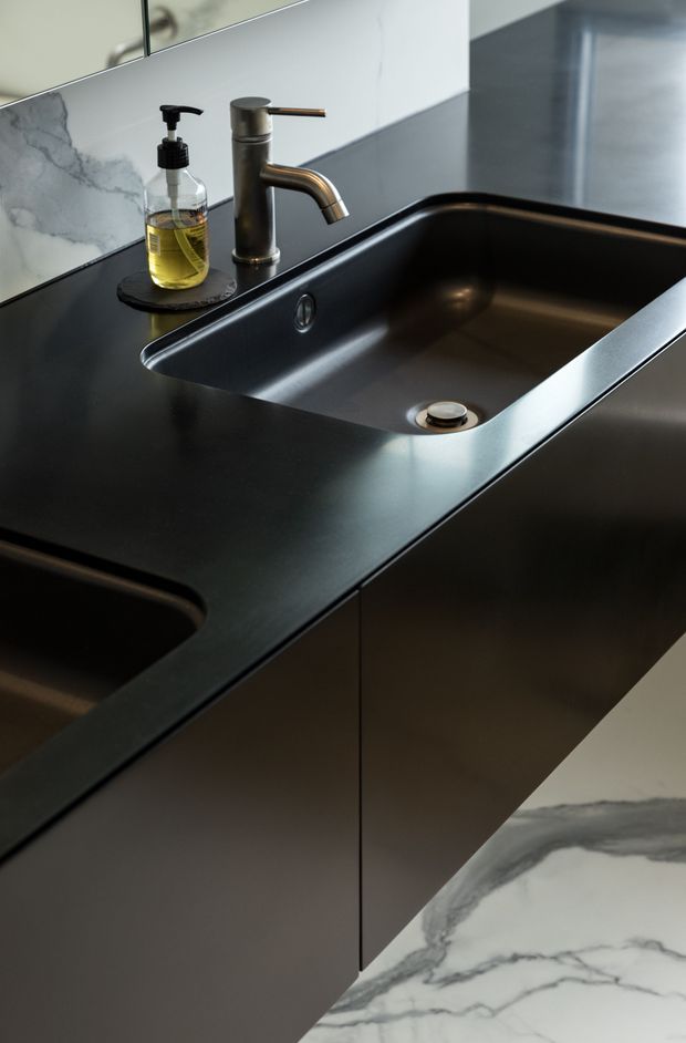 Close-up view of a refined bathroom vanity with a deep black integrated sink and dark countertop, paired with a brushed bronze single-handle tap. Dark flat-panel cabinetry sits below, while white marble-look wall and floor surfaces with grey veining provide contrast. A mirrored splashback reflects the surrounding space.
