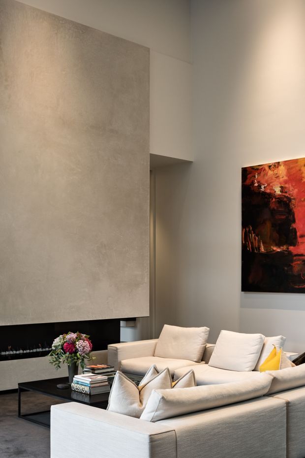 A refined contemporary living room with a large pale textured feature wall above a dark fireplace, warm neutral walls, a deep cream sectional sofa with multiple cushions, and a low black coffee table. An abstract red, orange, and black artwork adds a dramatic accent, while soft ambient lighting creates an elegant, inviting atmosphere.