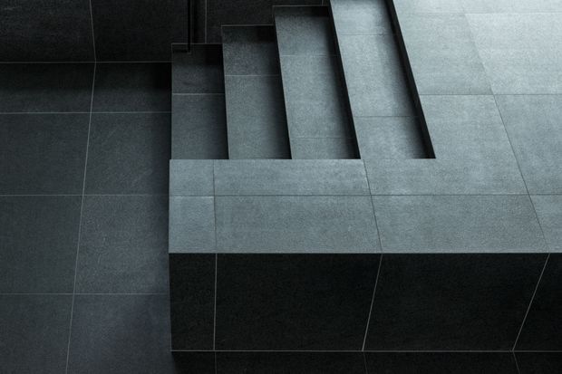 Close-up architectural view of a minimalist tiled stairway and landing. Large-format dark grey tiles cover the stair treads, risers, surrounding floor, and the projecting landing edge, creating a stark monochromatic composition with clean grout lines and strong geometric repetition.