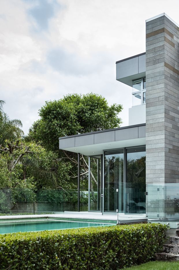 Exterior view of a contemporary multi-level residence with crisp flat rooflines, extensive floor-to-ceiling glazing, projecting balconies, and a tall vertically stacked masonry-clad feature wall. A swimming pool, glass balustrades, clipped hedges, and landscaped garden occupy the foreground.