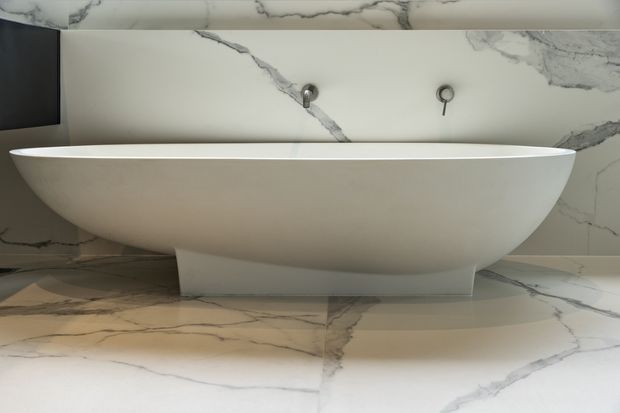 A close-up of a luxurious contemporary bathroom featuring a freestanding white oval bathtub on a pedestal base. Large-format white marble with prominent grey veining covers the wall and floor, with minimalist wall-mounted tap fixtures and a dark vertical panel at the left edge.