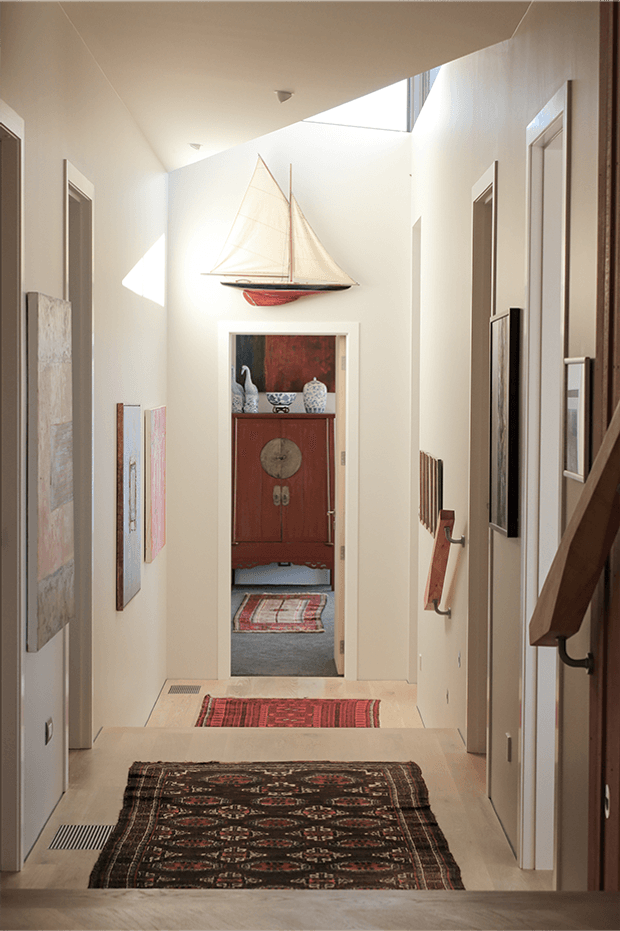 A bright, narrow residential hallway with cream-painted walls, pale timber flooring, multiple doorways, skylight-like clerestory glazing, and a timber stair handrail at right. Several patterned runners line the floor, while framed artwork, a wall-mounted model sailboat, and a red antique-style cabinet provide decorative focal points at the end of the corridor.