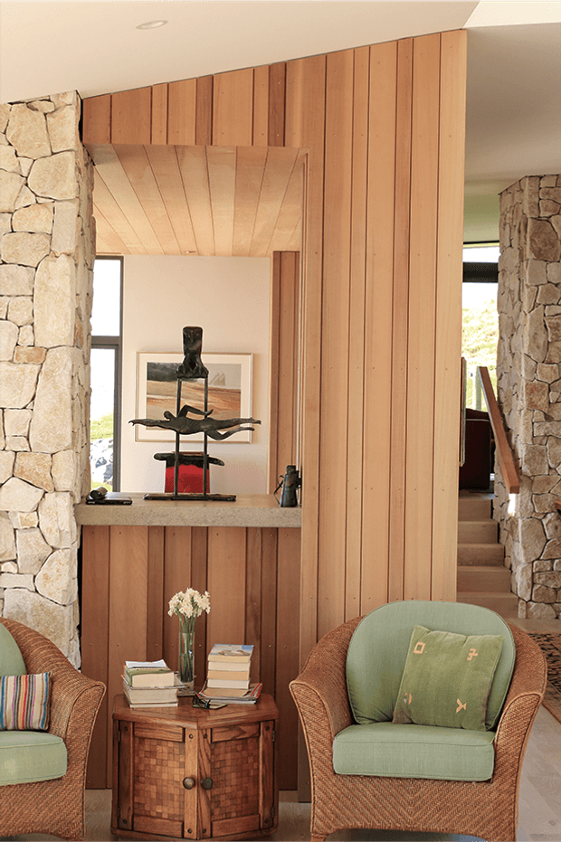 Warm, textural interior view of a residential entry or living area, framed by rough natural-stone walls and extensive vertical timber panelling. A timber-lined ceiling recess leads toward a small hall and staircase, while woven rattan armchairs with muted green cushions flank a carved timber side table. A stone-topped built-in console displays sculptural decor and artwork. The space has a refined rustic, nature-inspired character with strong material contrast between stone, timber and woven furniture.
