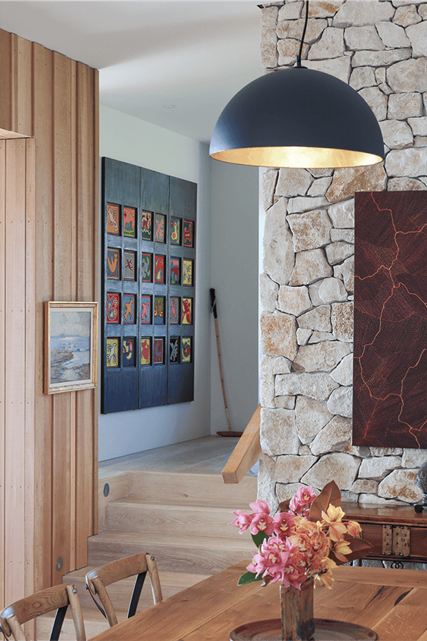 A warm, refined dining area opening into a raised hallway or living zone. The room features a substantial natural stone feature wall, vertical timber wall panelling, wide timber flooring, a large black dome pendant with a metallic interior, timber dining furniture, colourful framed artwork, and floral decoration. The composition highlights layered levels and handcrafted natural materials.