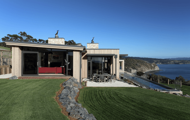 Exterior view of a low-slung modern residence on a landscaped sloping site, with strong horizontal massing, pale vertical cladding, large glazed openings, flat roof sections, and two stone-clad chimney elements. A furnished outdoor dining area sits beside the house, with lawn and a rugged stone drainage or retaining feature in the foreground. The property overlooks a dramatic coastal inlet and headland.