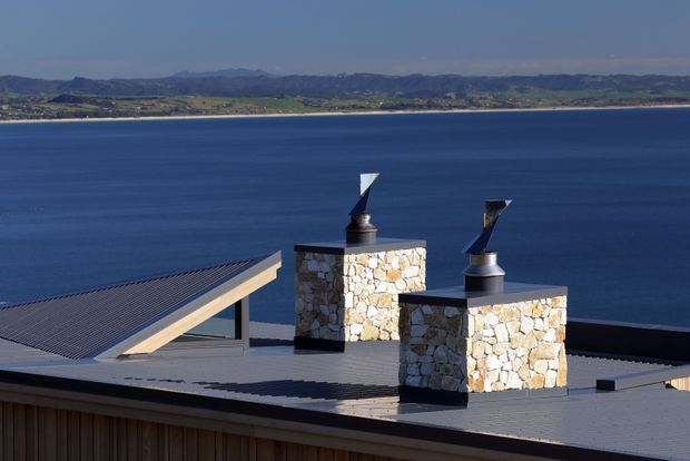 Exterior architectural view focusing on a modern low-pitched roof beside a large body of water. The roof has two substantial stone-clad chimney structures with dark metal caps and angular wind deflectors, plus a sloped glazed roof element at left. Distant hills and shoreline form the background. The palette is dominated by dark grey roofing, pale natural stone, timber-toned cladding, and deep blue water.