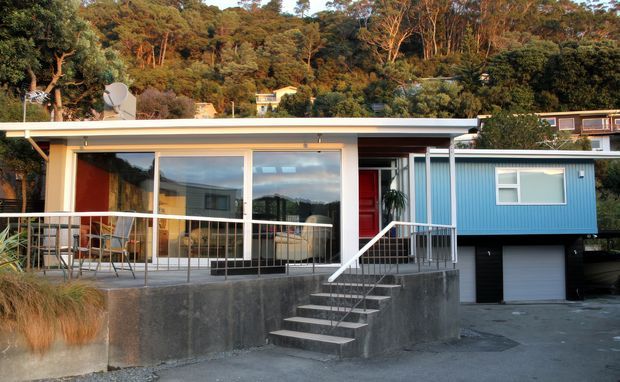 Single-storey modern residence set into a wooded hillside, with a low-pitched flat roof, extensive floor-to-ceiling glazing, a covered terrace, and a distinctive blue corrugated-cladding wing above an integrated garage. Concrete retaining walls and external steps define the elevated entry, while mature bushland surrounds the property.