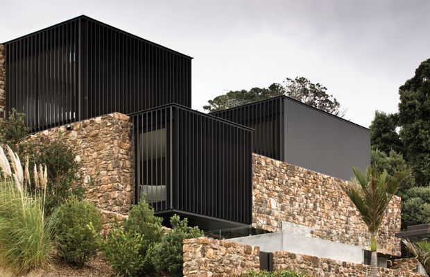 A contemporary multi-level building set into a landscaped sloping site. The exterior combines dark vertical ribbed metal cladding and dark metal-framed glazing with extensive rough natural stone retaining walls and masonry. Layered flat-roofed volumes step up the hillside, surrounded by grasses, shrubs, and trees under an overcast sky.