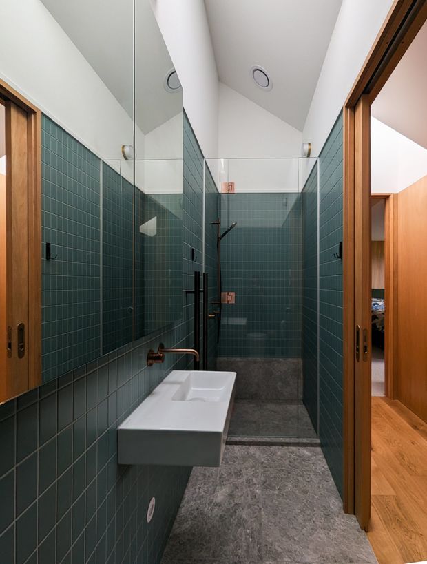 A narrow contemporary bathroom with deep teal-green square tiled walls, a wall-mounted white rectangular basin, exposed copper-toned tapware, and a glass-enclosed walk-in shower at the rear. Grey stone-look flooring, timber-framed sliding doors, recessed ceiling lights, and a timber-lined adjoining space visible through the doorway create a refined, modern interior.