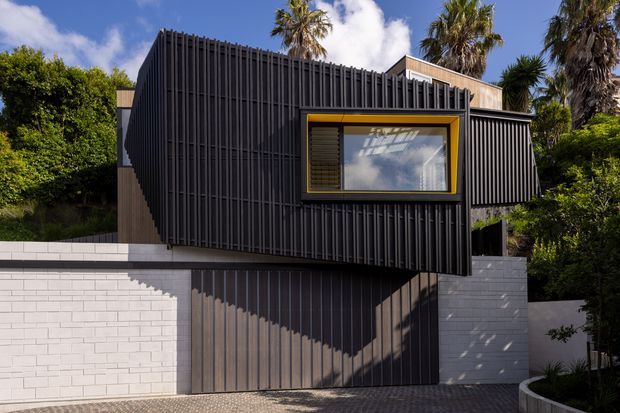 Contemporary multi-level residential facade with dark vertical ribbed cladding, a projecting upper volume, and a distinctive mustard-yellow framed window. A dark garage or entry door sits below, flanked by white masonry walls, with tropical landscaping and palm trees around the property.