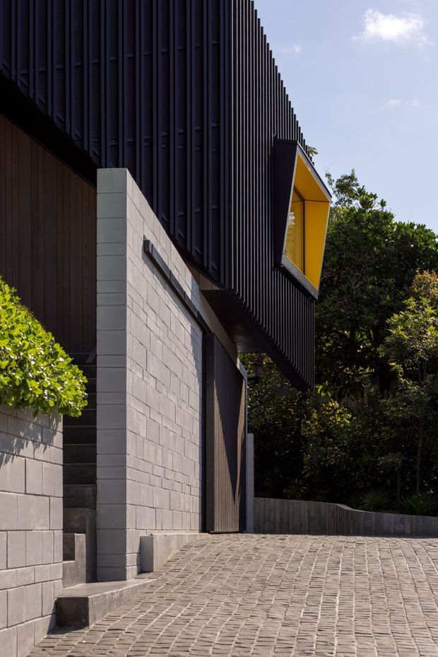 A close exterior view of a contemporary multi-level building on a landscaped, sloping site. The facade combines dark vertical metal fins and timber-toned cladding with pale concrete block walls, a projecting angular box with a vivid yellow recessed underside, and a broad cobbled driveway or paved forecourt. Dense planting surrounds the structure.