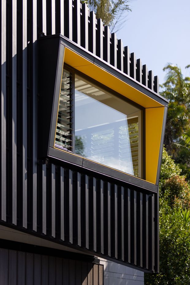 Close-up exterior view of a contemporary building facade clad in vertically ribbed black panels. A prominent projecting window is framed in black with a vivid yellow recessed reveal, with louvred glazing visible at the side and reflections of the surrounding landscape in the glass. Vegetation and blue sky appear in the background.