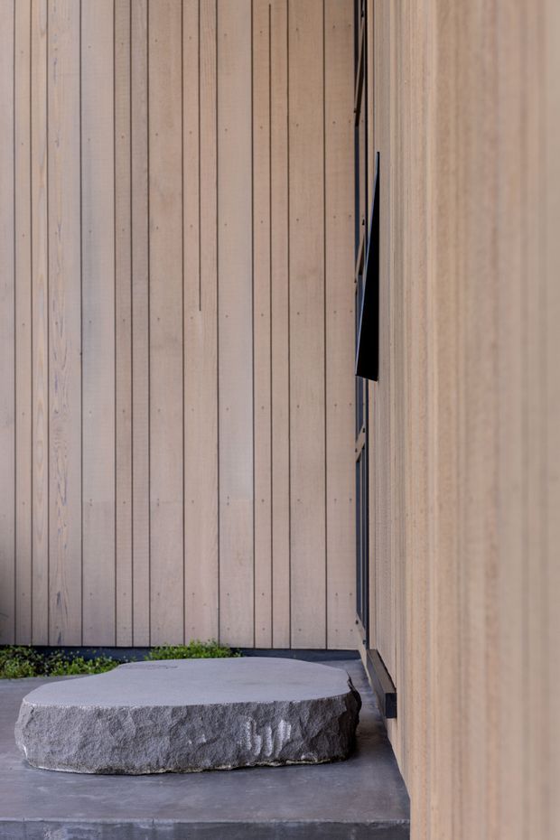 A narrow contemporary exterior passage or entry court lined with vertical pale timber cladding. A rough-hewn circular natural stone slab sits on a smooth grey concrete platform, with a slim dark-framed opening and small strip of planted greenery adding contrast. The composition is minimalist and tactile, with strong vertical lines and muted natural materials.