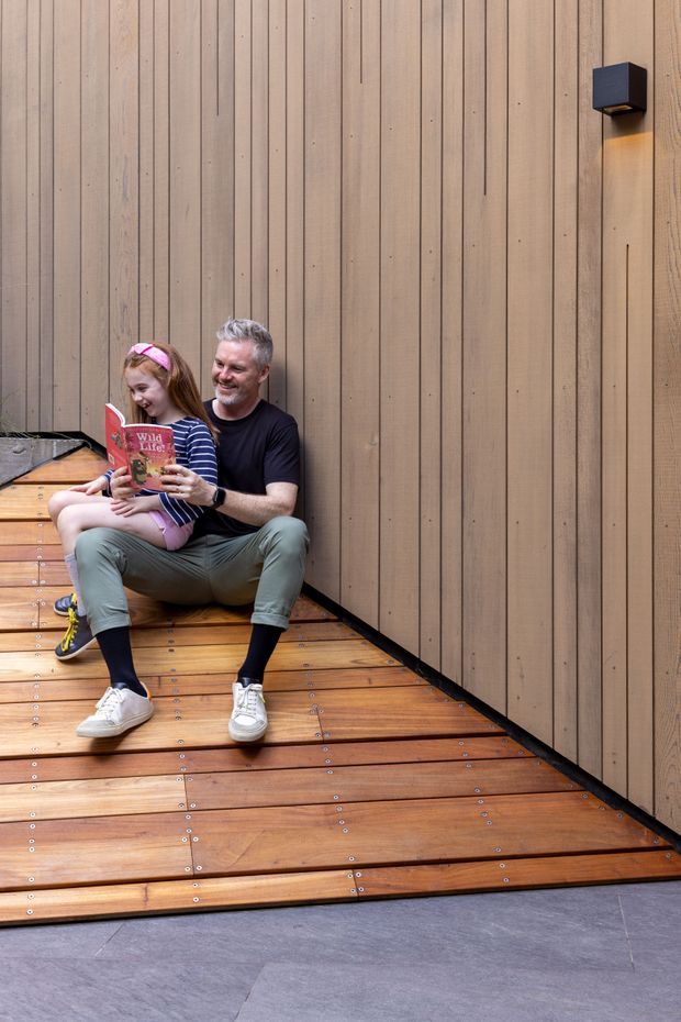 A warm, contemporary timber-lined interior or covered outdoor nook with vertical natural wood wall panelling and a raised horizontal timber platform floor secured with visible metal fasteners. A father and child sit reading together on the steps, while a compact dark wall light provides a subtle accent. Grey stone or concrete paving appears in the foreground.