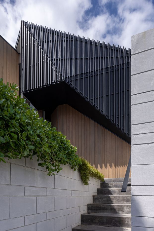 Exterior architectural photograph of a modern multi-level residence, focused on a stairway leading toward a dark-clad upper volume. The facade combines vertical dark metal screening and battens, warm vertical timber wall cladding, and pale rendered or concrete block walls. Strong geometric lines, cantilevered construction, and landscaped planting frame the entry stairs beneath a partly cloudy sky.