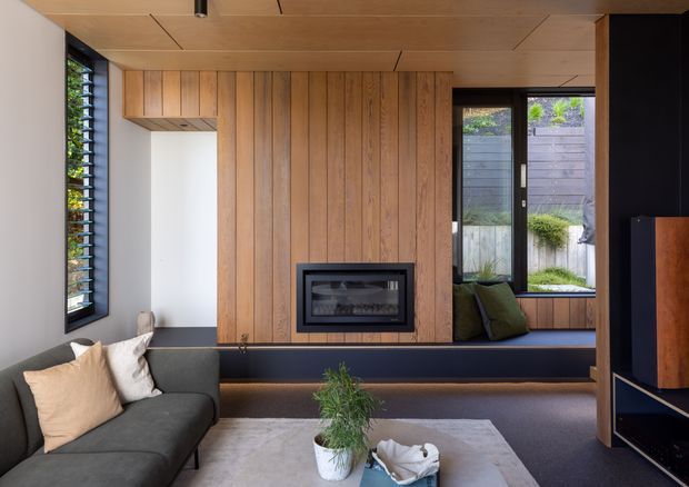 Contemporary living room with a dark sectional sofa, built-in window seat, timber wall panelling and integrated fireplace. The room features a warm timber-lined ceiling and joinery, black-framed windows, dark flooring, and a neutral textured rug with restrained furnishings. A landscaped courtyard is visible through the glazing.
