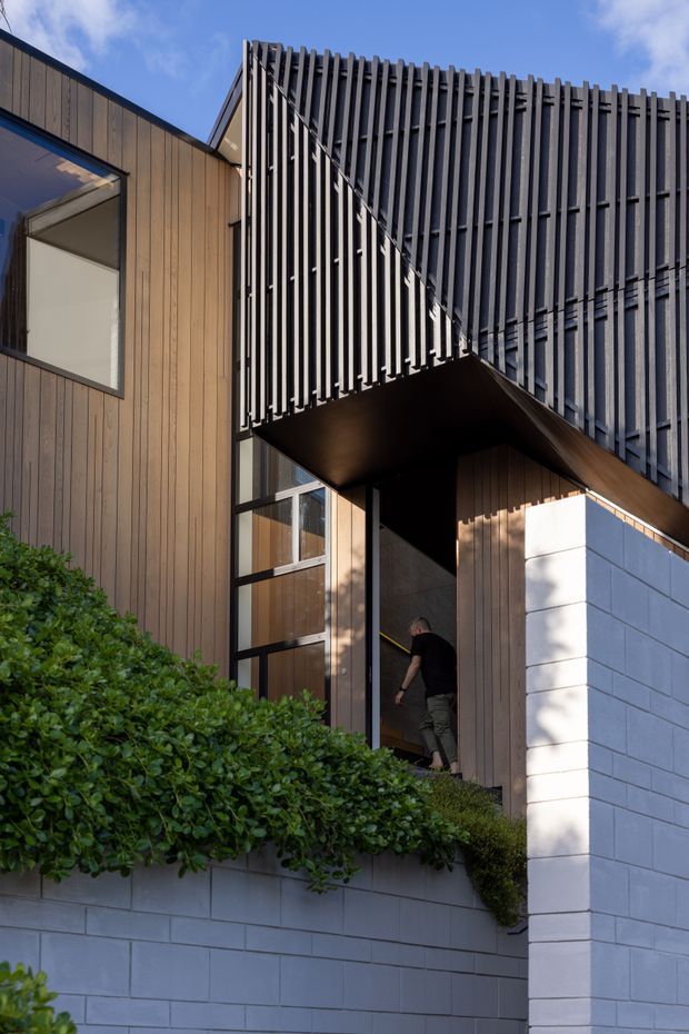 Exterior architectural photograph of a contemporary multi-level home, focused on the entrance and projecting upper volume. The facade combines vertical dark timber cladding, a deep charcoal-grey vertical screen or fin facade, black-framed glazing, and white painted masonry block walls. Dense hedging partially conceals the lower level, while strong sunlight creates defined shadows across the facade.