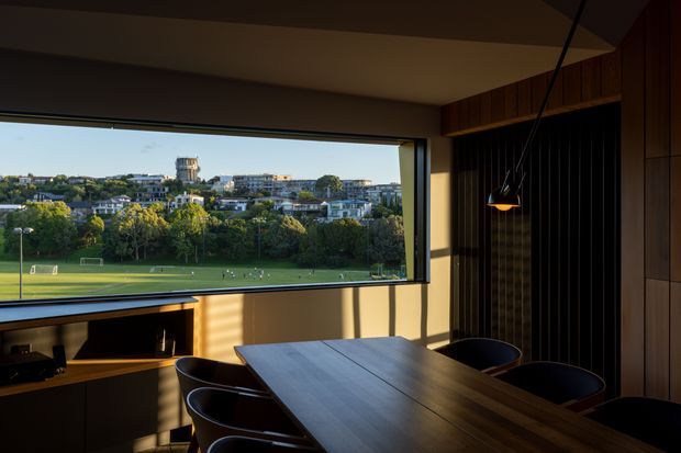 A warm, dimly lit contemporary dining room with a large picture window overlooking a green sports field and suburban cityscape. The room features a long dark timber dining table with several black sculptural chairs, dark timber wall panelling and built-in cabinetry, a slim black pendant light, and a low media or storage unit beneath the window. Strong late-afternoon sunlight creates dramatic shadows across the interior.