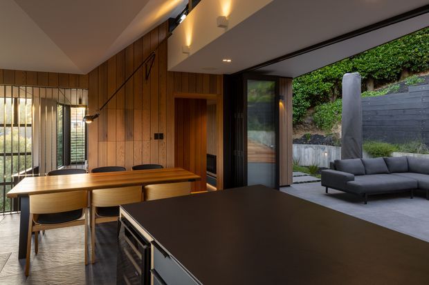 A refined open-plan dining and kitchen interior with a warm timber-panelled feature wall, long timber dining table, dark kitchen island, and large glazed openings connecting to a landscaped outdoor courtyard. Slim black-framed glazing, integrated lighting, and contemporary built-in elements create a calm, modern indoor-outdoor setting.