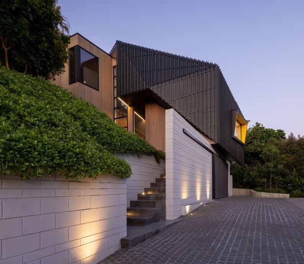 Twilight exterior view of a contemporary multi-level residence on a sloping site. The house features warm timber cladding, a prominent curved or angled facade screened with closely spaced dark vertical battens, large glazed openings, a white masonry garage and retaining wall, illuminated external stairs, and a paved driveway. Dense landscaped planting frames the building.