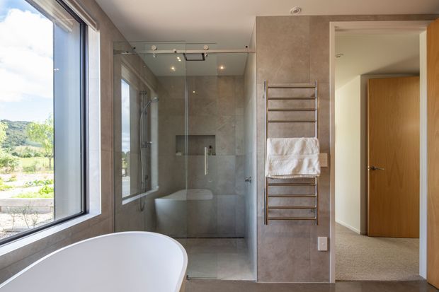 Contemporary bathroom with a freestanding white bathtub in the foreground, a glass-enclosed walk-in shower, large-format grey stone-look wall and floor finishes, and a heated towel rail. Full-height glazing provides a bright outlook to the landscape, while a timber door opens toward an adjoining carpeted space.