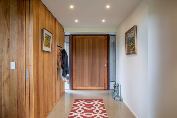A contemporary residential entry hallway with extensive warm timber wall panelling and matching built-in storage on the left, a large timber front door framed by grey stone-like panels, and pale polished flooring. The space includes recessed ceiling downlights, coat hooks with hanging garments, framed artwork, decorative objects, and a red-and-cream patterned runner. The corridor is bright, clean, and warmly finished.