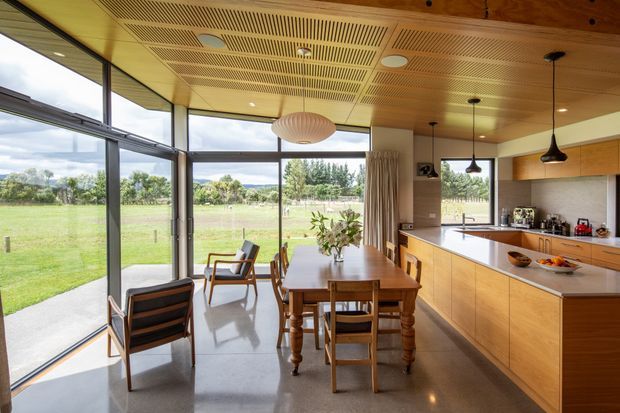 A bright open-plan kitchen and dining area with expansive floor-to-ceiling glazing overlooking a rural landscape. The room features warm timber cabinetry and a timber slatted acoustic ceiling, a polished grey concrete floor, a long island with integrated cabinetry, pendant lighting, and a timber dining table with chairs. Lounge chairs sit beside the glazing, creating a connection to the outdoors.