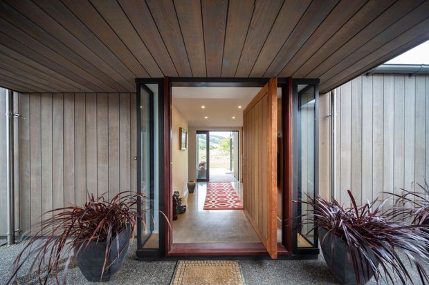 A covered residential entryway viewed from outside, framed by dark timber soffit and vertical weatherboard walls. Glazed sidelights flank a timber front door, opening to a long interior hallway with recessed ceiling lights, a patterned runner, and a view through to the landscape beyond. Large planters with burgundy foliage sit on either side of the entrance.