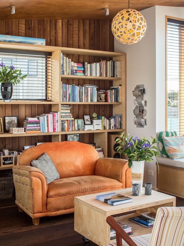 A warm, richly layered living room with a built-in timber bookcase covering a dark vertical wood-panel feature wall. The room includes a large cognac leather sofa, a light timber coffee table, abundant books, potted plants, framed objects, and a spherical sculptural pendant light. Timber-framed windows with horizontal blinds bring in daylight, while a window seat with patterned cushions is visible at right. The overall look is eclectic, organic, and inviting, with strong mid-century influences.