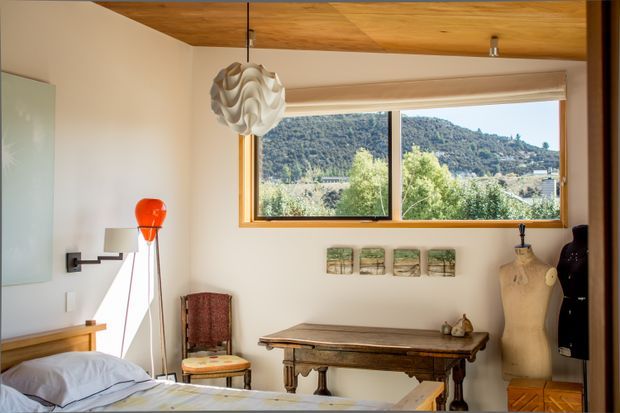 A warm, compact bedroom with a bed in the foreground, a rustic timber console or work table, vintage chair, mannequin, and sculptural pendant light. A large timber-framed window provides a view of a wooded hillside and valley. The room has white walls, a timber-lined ceiling, and eclectic vintage furnishings.