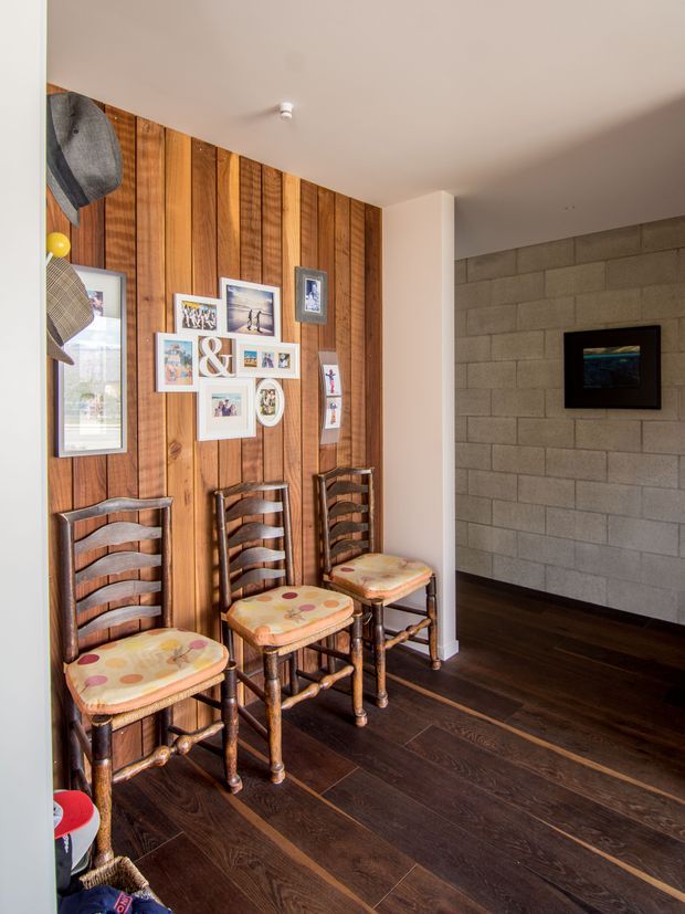 A residential interior hallway or entry area featuring three vintage-style wooden ladder-back chairs with patterned upholstered seats arranged against a vertical timber feature wall. Framed family photographs and artwork are displayed above the chairs. The adjacent wall is finished in grey concrete block, with a small wall-mounted screen or artwork. The room has dark timber flooring, white plasterboard ceilings and trim, and warm natural light.