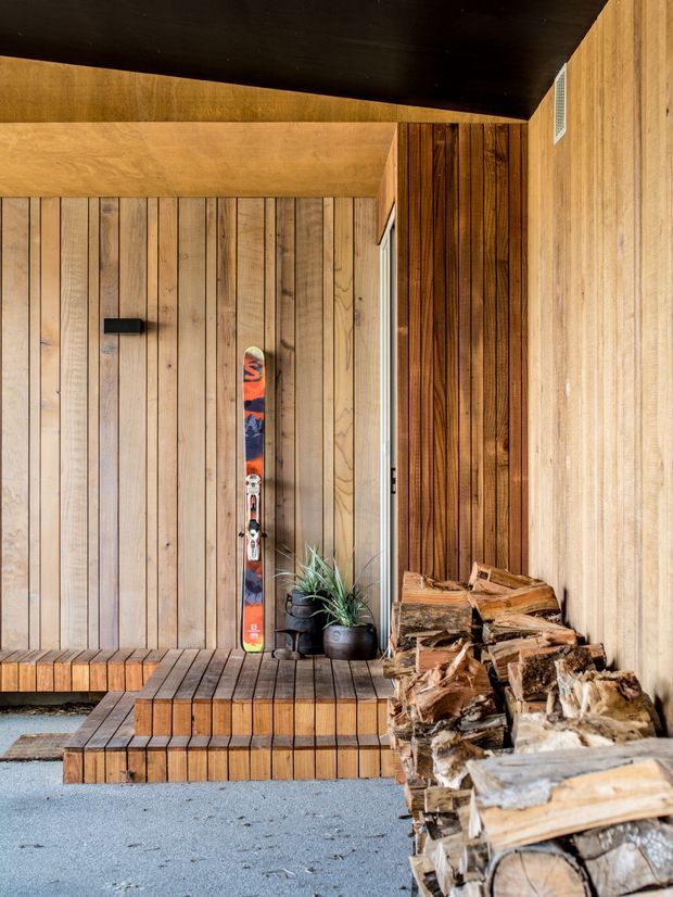 Warm, timber-lined entry or mudroom-style space featuring a built-in slatted timber bench and stepped platform, vertical timber wall panelling, a mounted ski, potted plants, and a substantial stack of firewood. The predominantly wood-clad interior has a rustic alpine character and subdued natural lighting.