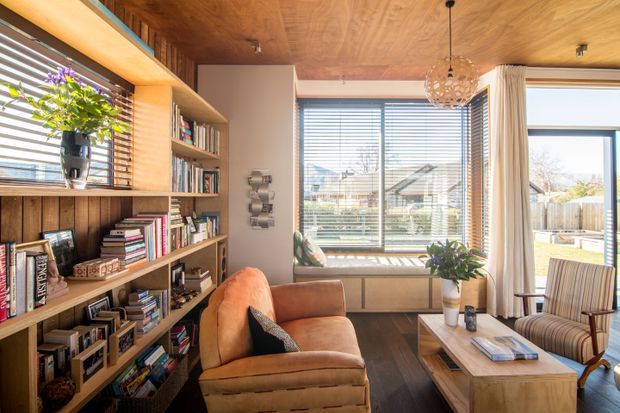 Warm, light-filled living room with extensive built-in timber shelving filled with books, a caramel leather armchair, a striped upholstered lounge chair, and a timber coffee table. The room features a wood-panelled ceiling, dark timber flooring, large windows with horizontal blinds, cream curtains, and a sculptural pendant light. A small outdoor area and neighbouring homes are visible through the glazing.
