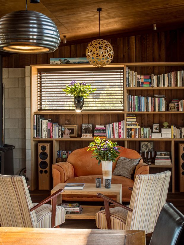 A warm, richly detailed living room with dark timber wall and ceiling panelling, full-height built-in bookshelves, a central window with timber venetian blinds, a tan leather sofa, striped upholstered lounge chairs, and vintage-style pendant lighting. A timber coffee table, books, framed photographs, speakers, and floral arrangements create a cozy library-like atmosphere.