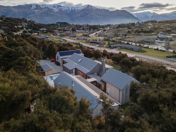 Aerial view of a contemporary multi-wing house with grey metal pitched roofs and vertically clad walls, nestled among dense native vegetation on a sloping site. The surrounding suburban development, roads, and dramatic snow-dusted mountain ranges are clearly visible in the background.