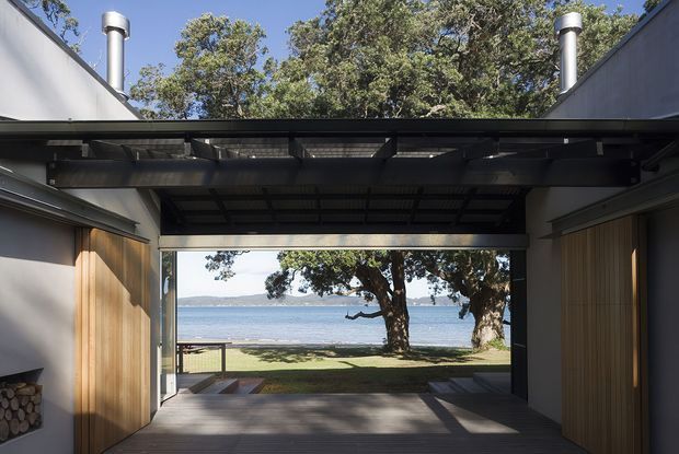 A dramatic open-sided architectural pavilion or passage frames a waterfront view. Dark steel roof beams and translucent roof panels span between pale walls, with warm vertical timber cladding on both sides, exposed cylindrical flues, and a timber deck leading toward a lawn, trees, and calm water beyond. The composition emphasizes indoor–outdoor connection and strong horizontal framing.
