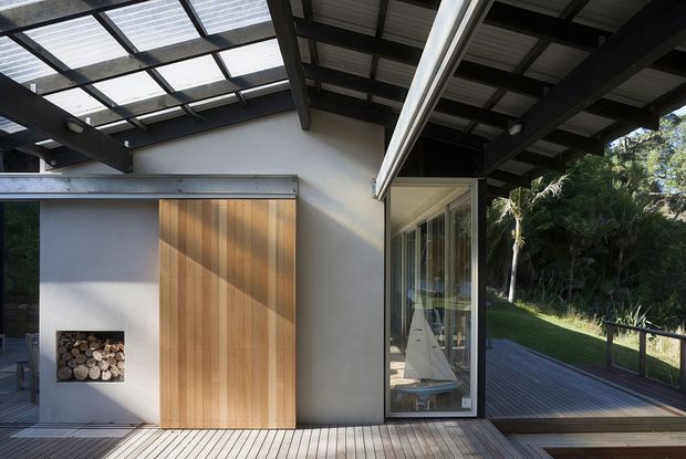 Contemporary residential exterior or covered outdoor living area with a timber deck, exposed dark structural beams, translucent roof panels, and a pale rendered wall. A tall timber sliding screen conceals a recessed outdoor fireplace, while full-height glazing reveals a sailboat-themed interior detail. The deck extends toward a lush landscaped setting with a simple dark balustrade.