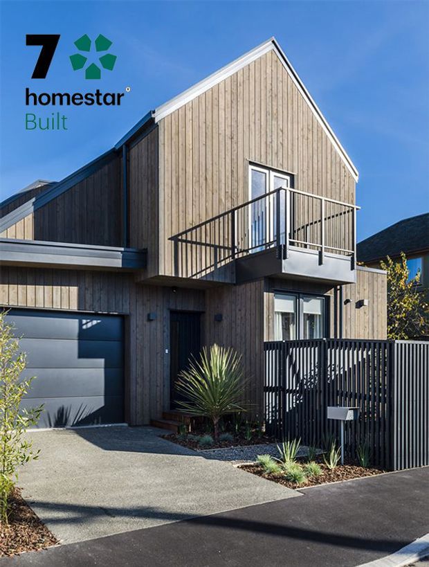 Exterior photograph of a contemporary multi-level townhouse with a steep gable roof and vertical timber-clad facade. The home features a projecting upper-floor balcony with black metal balustrade, dark-framed glazing, an integrated garage, black vertical-slatted fencing, and a paved driveway with drought-tolerant planting. Bright clear daylight emphasizes the natural timber and dark contrasting details.
