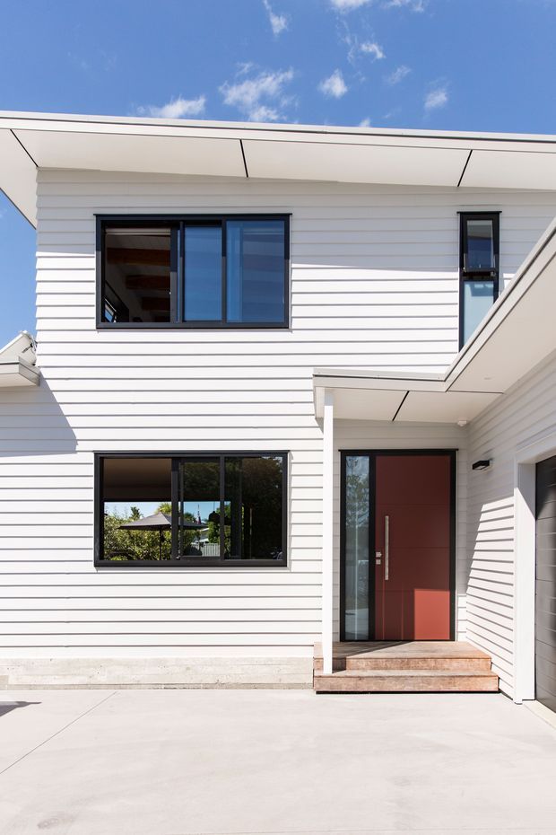 A bright exterior view of a contemporary two-storey house with white horizontal weatherboard cladding, black-framed windows, a deep roof overhang, and a recessed entry porch. The front door is a muted red-brown, reached by timber steps, with a concrete driveway in the foreground and blue sky above.