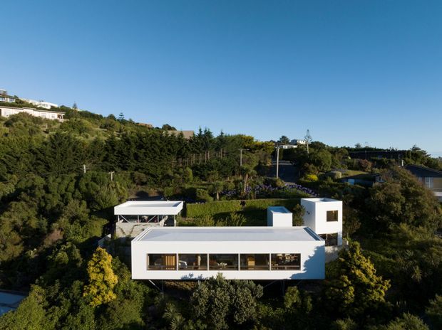 Aerial view of a contemporary white residential house set among dense mature landscaping on a sloping suburban site. The main building has crisp geometric volumes, a flat roof, extensive horizontal glazing, and a detached or adjoining flat-roofed pavilion visible behind it. Other hillside residences and garden vegetation surround the property.