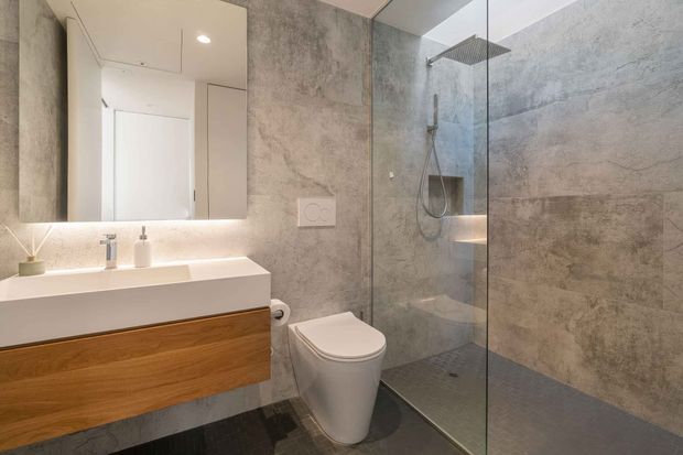 Contemporary bathroom with a floating warm timber vanity, white rectangular basin, wall-mounted fixtures, mirrored cabinet, wall-hung toilet, and a frameless glass walk-in shower. Large grey stone-look wall tiles and dark floor tiles create a clean, refined finish.