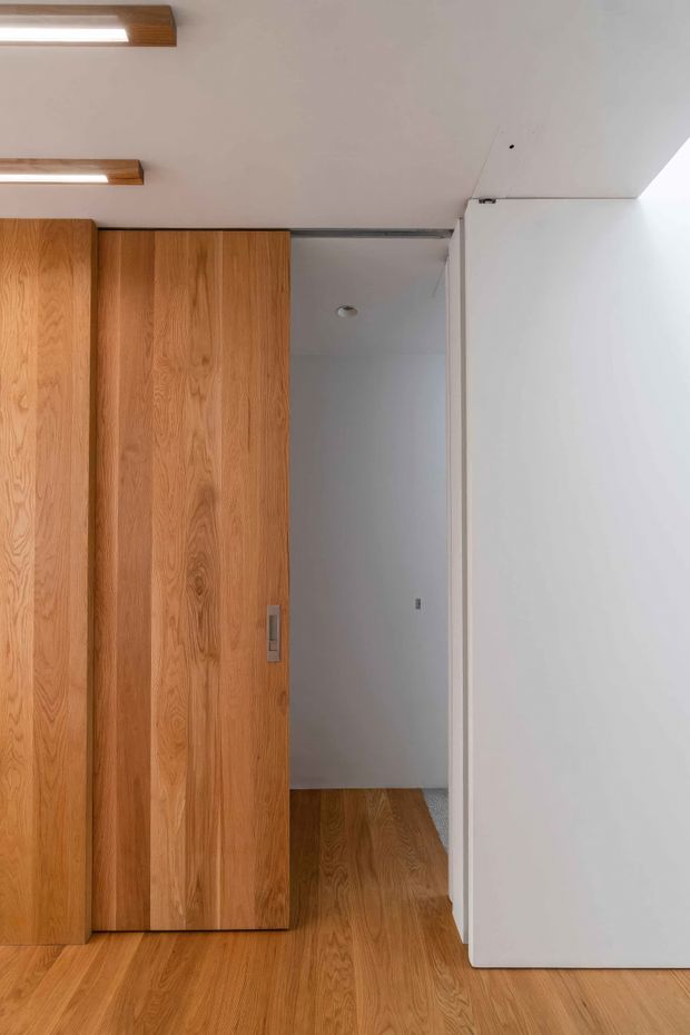 Minimal interior view of a warm timber-lined sliding door or built-in partition opening into a small white room or hallway. The space features light oak timber flooring, matching vertical timber panels, white plasterboard walls and ceiling, recessed lighting, and slim linear timber-and-white ceiling fixtures. The composition is clean and understated, with strong emphasis on built-in joinery and material continuity.