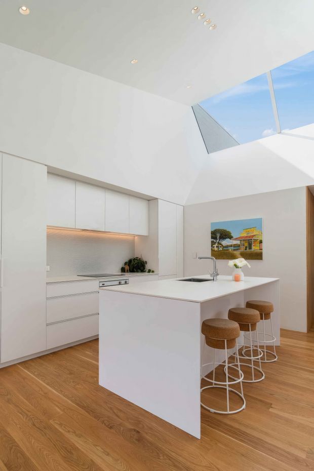 Bright contemporary kitchen with a large white island, integrated white cabinetry, concealed rangehood, cooktop, sink and four bar stools. Warm timber flooring contrasts with the white surfaces, while recessed ceiling lights and a large overhead skylight bring in abundant natural light. A colourful artwork is displayed on the wall beside the kitchen.
