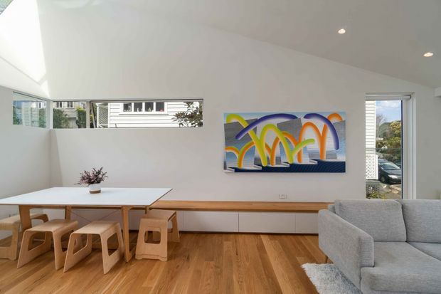Bright, contemporary open-plan living and dining area with white walls, warm timber flooring, a built-in timber media cabinet, pale wood dining table and stools, and a light grey sofa. Narrow horizontal windows and a tall glazed opening bring in natural light, while a colourful abstract artwork provides a vivid focal point.
