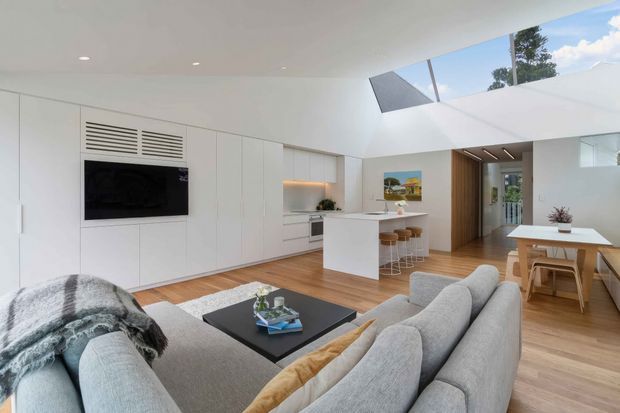Bright open-plan contemporary living, dining and kitchen area with white full-height built-in joinery, integrated television, timber flooring, a central kitchen island with bar stools, skylights, and a grey sectional sofa. A timber-lined doorway leads toward another part of the home.
